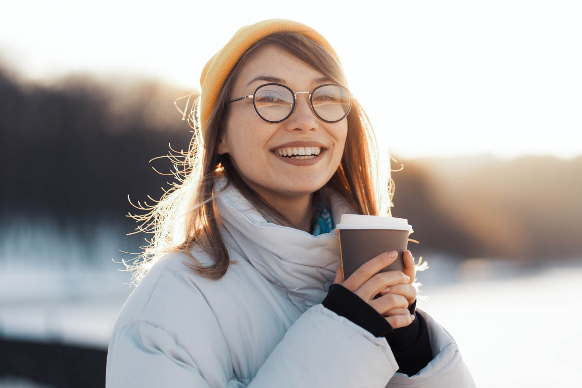 A woman is wrapped up in winter clothes and holding a hot drink outside