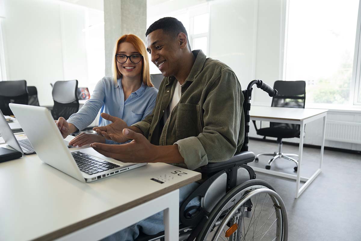 A man in a wheelchair sitting next to a woman at a desk, both individuals are in conversation as they look at a laptop screen