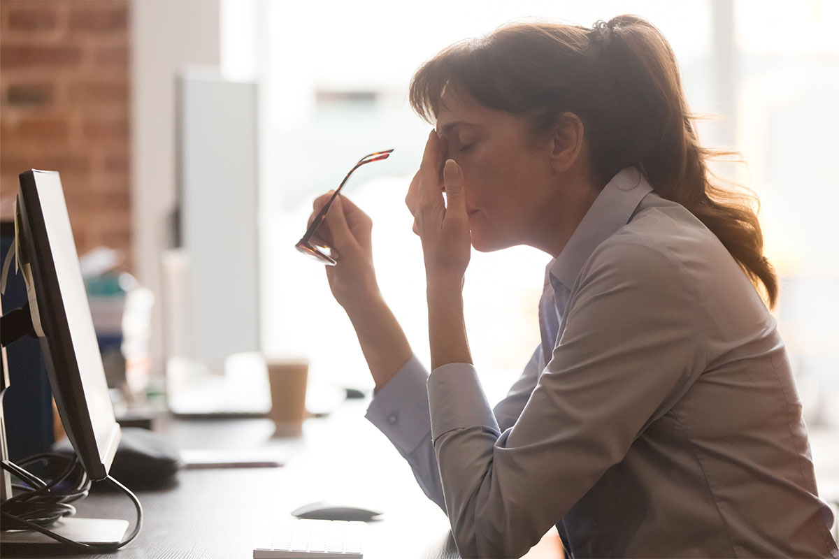 A woman sits at her desk looking burntout. She is touching her head as if she has a headache.