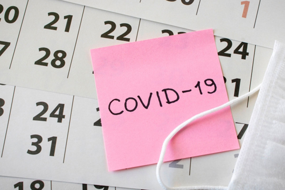 Pages of a calendar are spread out. On top of the pages sits a pink post it reading 'COVID-19' and a PPE face mask.