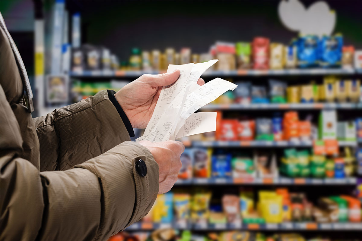 Against the background of stacked supermarket shelves, a pair of hands holds a stack of receipts.