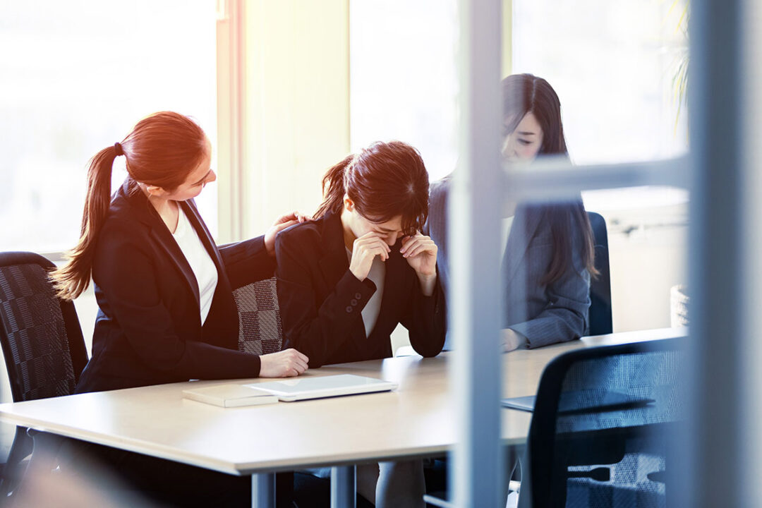 Three businesswomen sit at a desk in an office. The middle woman is crying as the two women either side of her comfort her.