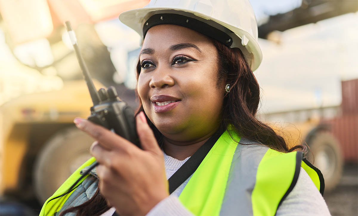 A black woman with a walkie talkie, hard hat and high-vis jacket