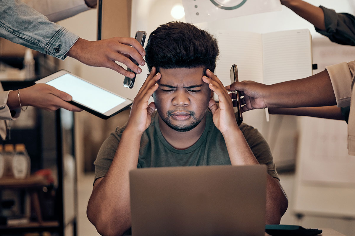 A man holds his head in his hands as he looks distressed in front of his laptop. He is surrounded by hands holding devices such as phones and tablets.